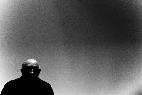 Portrait noir et blanc à contre-jour de Raymond Depardon, en bas à gauche,  sur fond gris